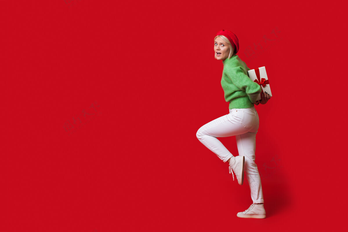 广告一位金发白种女人背着一个礼物盒 戴着一顶帽子 看着一面红墙上的相机 还有空余的空间年轻空白销售
