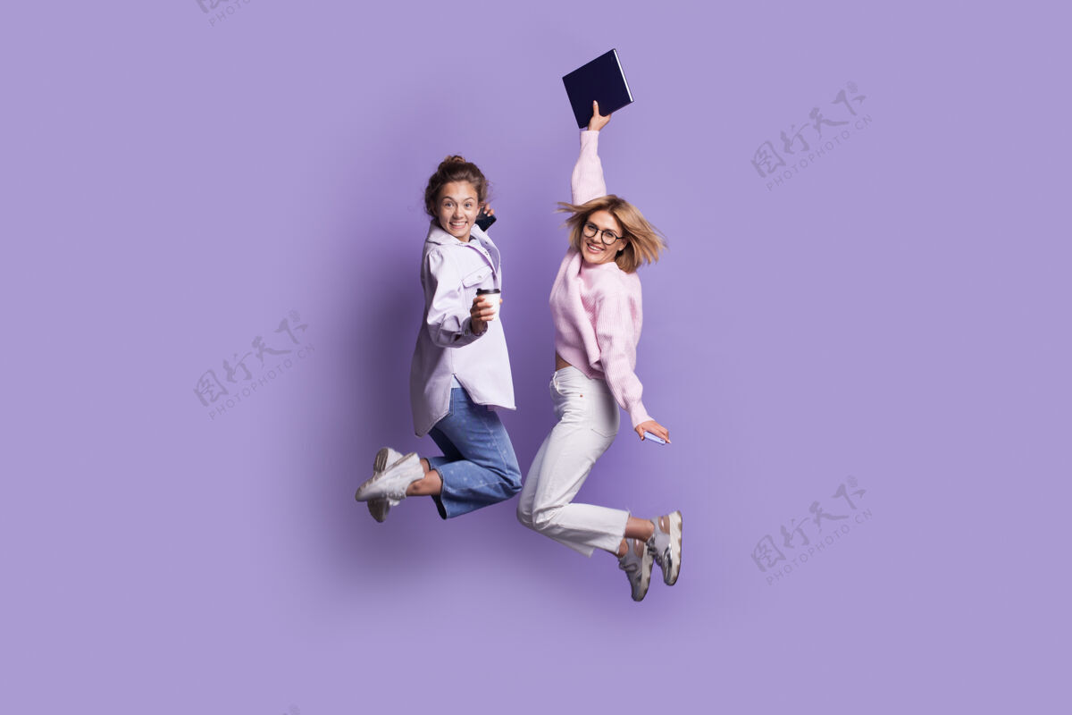 微笑两个穿着休闲服的高加索学生拿着一本书 对着镜头微笑着跳上了紫罗兰色的画室墙上女孩跳跃兴奋