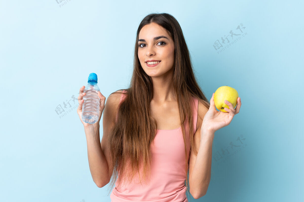 矿物质一个年轻的白人妇女 拿着一个苹果和一瓶水 被隔离在蓝色的背景下女性五颜六色瓶子