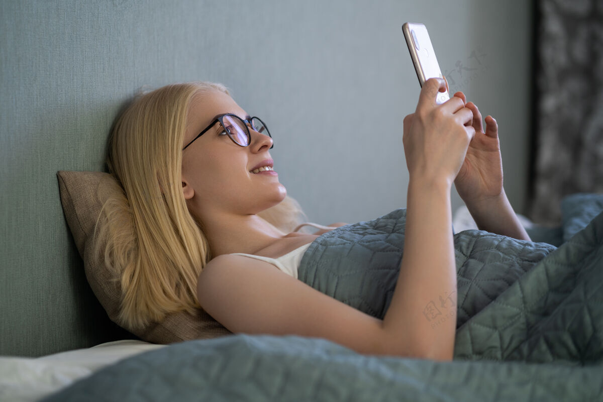 休息微笑的女人看着手机躺在白色的床上床快乐金发碧眼的年轻女子在家里用手机家漂亮女孩在智能手机上打字和上网打字年轻西班牙裔
