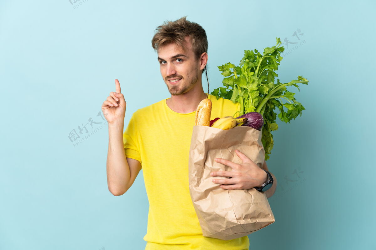 素食一个金发碧眼的人拿着一袋外卖食品 在蓝色的舞台上展示 举起一个手指表示最好人男性营养