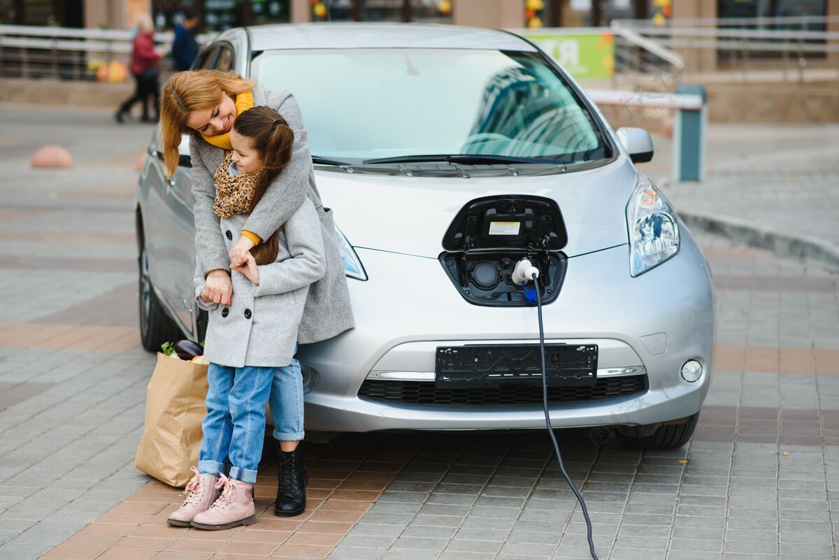 替代妈妈带着女儿在电动加油站给电动车充电 用手机说话选择街道充电