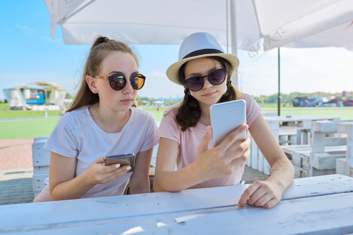 女性两个十几岁的女孩拿着智能手机坐在户外聊天青年咖啡馆 青少年 友谊 沟通 人的观念年轻微笑乐趣