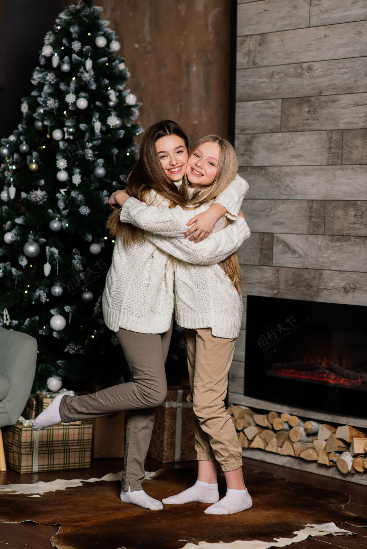 牵手两个可爱的女孩在圣诞节摆出亲吻的表情树长-站在树旁的一位留着头发的欧洲女士和礼物孩子姐妹国际