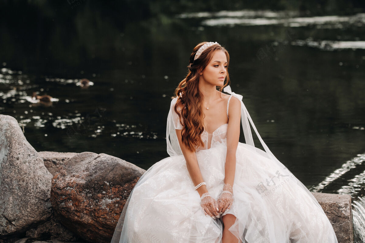 婚纱一位穿着白色礼服 戴着手套 赤脚的优雅新娘正坐在公园的瀑布旁 享受着大自然金发新娘手套