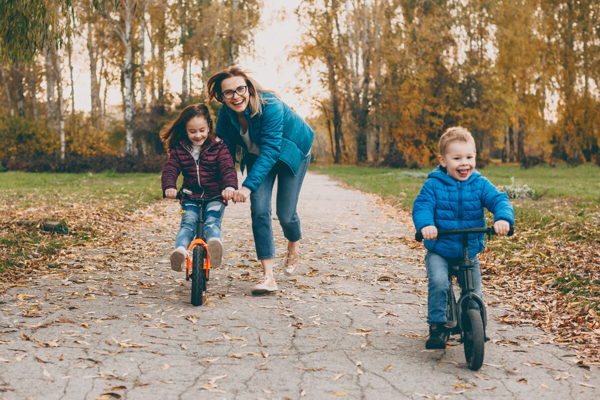 情感可爱的年轻妈妈在男孩走的时候和她的女儿和儿子一起骑自行车妈妈帮她的小女孩骑自行车秋天朋友妈妈