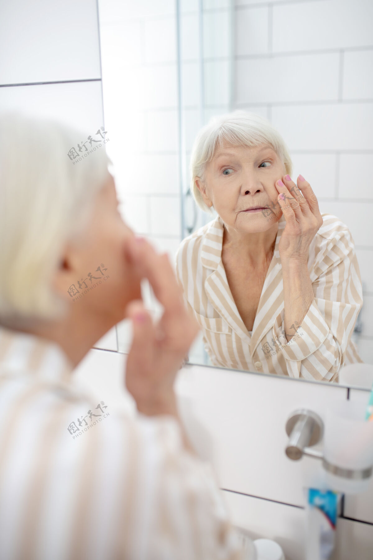 养老金老化灰色-一个满头头发的女人在浴室照镜子 抚摸她的脸清洁积极的老年人认证
