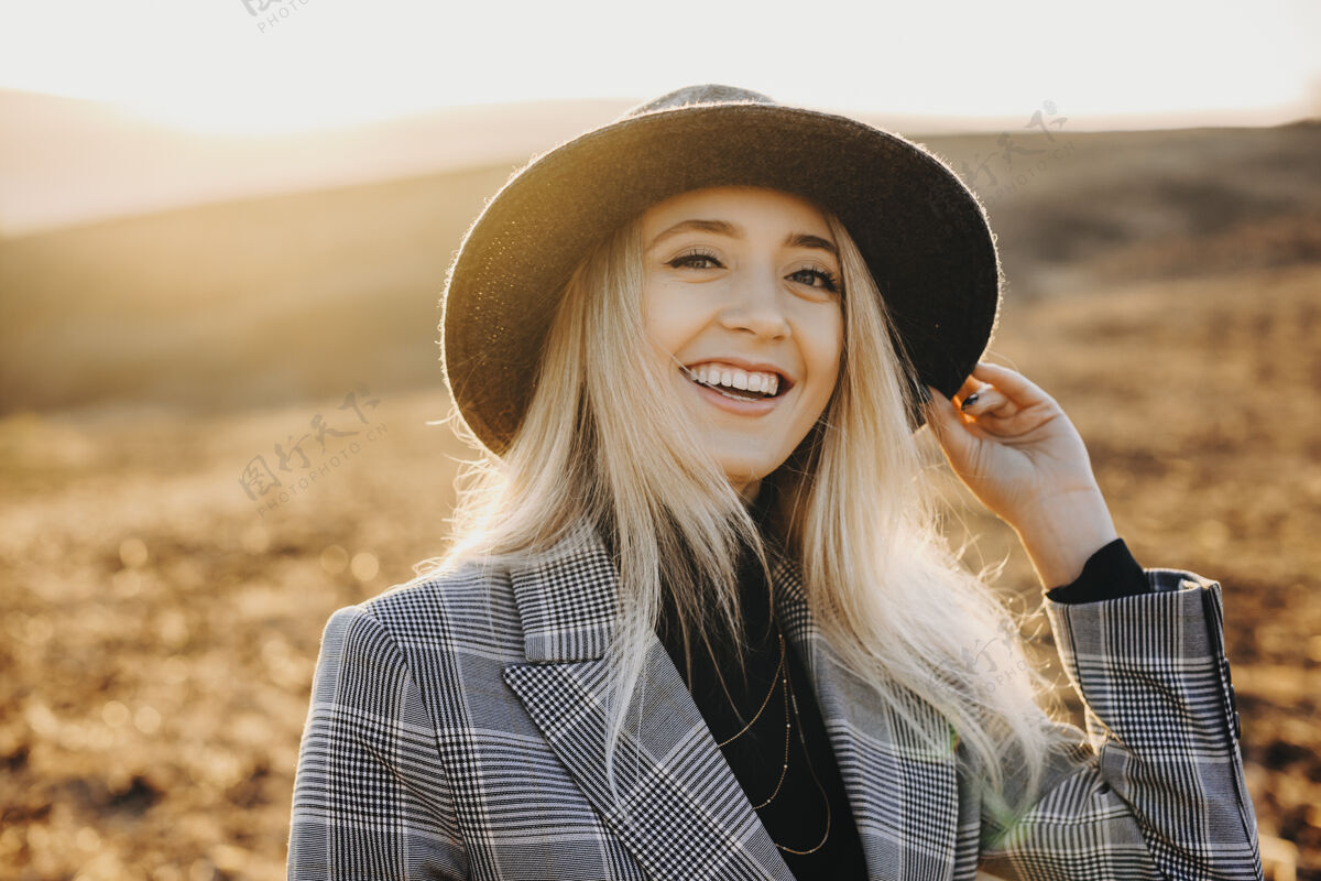 阳光一个可爱的年轻女孩笑着看着相机的肖像 一只手放在帽子上 顶着太阳升起 在假期里探索新的地方年轻肖像女性