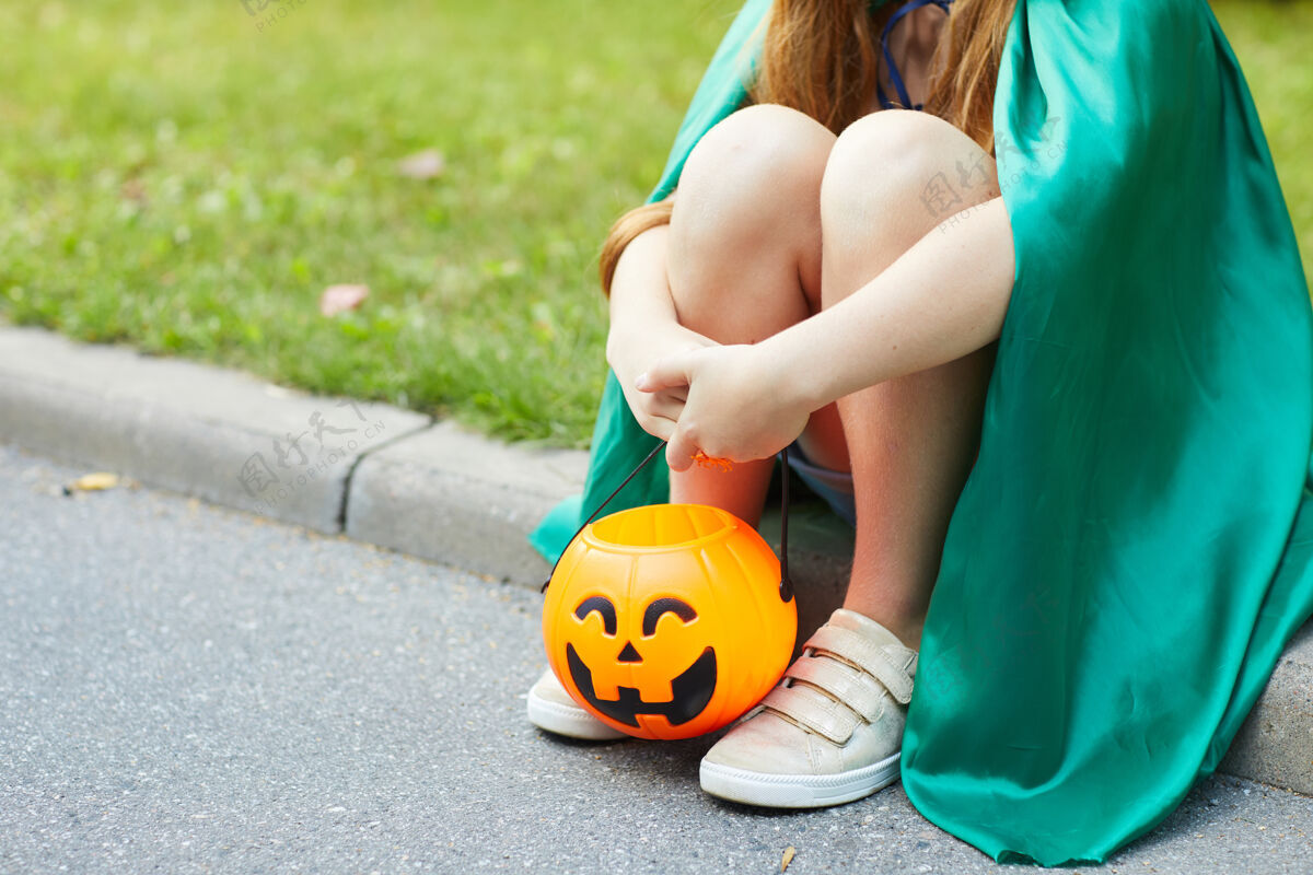 天特写镜头：穿着万圣节服装的小女孩坐在户外的地上 手里拿着南瓜篮装糖果桶街道享受