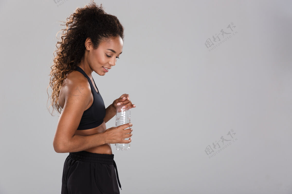 看一位年轻漂亮的非洲女运动员在灰色墙壁上独自训练后休息的肖像 手里拿着装满水的瓶子瓶子人美国
