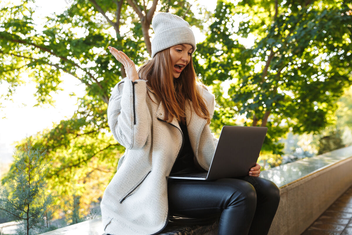 坐着美丽的年轻女子身着秋衣 戴着秋帽 坐在户外 用笔记本电脑 庆祝成功户外城市女性