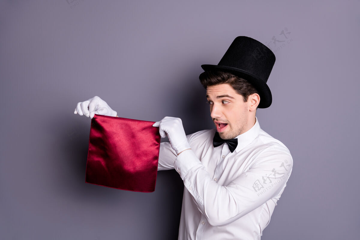 胡茬有趣的时髦魔术师开始他的精彩焦点牵手红色餐巾看说abracadabra穿白色衬衫黑色蝴蝶结帽子隔离灰色墙壁魔术娱乐表演