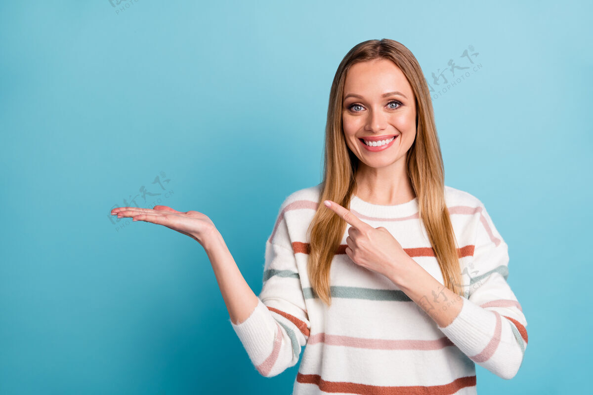 选择照片中开朗积极的美丽甜美的女人指着她手掌上的空白微笑着牙齿隔离粉彩蓝色墙壁休闲方式指标