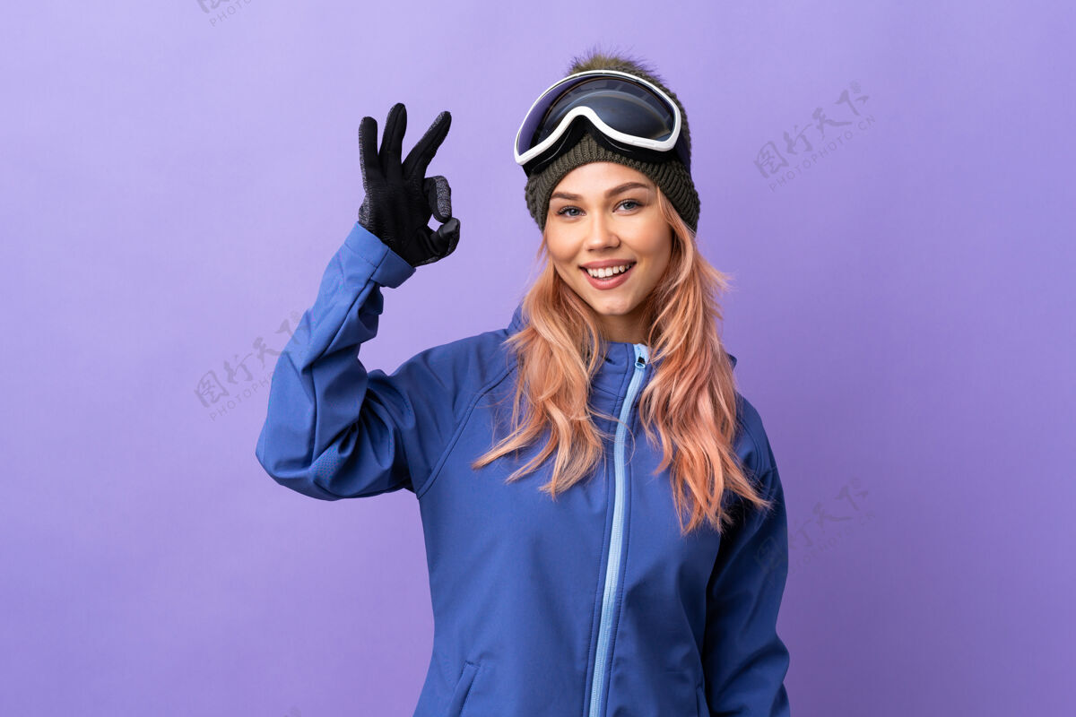 滑雪滑雪者女孩戴着滑雪板眼镜在孤立的紫色上用手指显示ok标志年轻女人玻璃