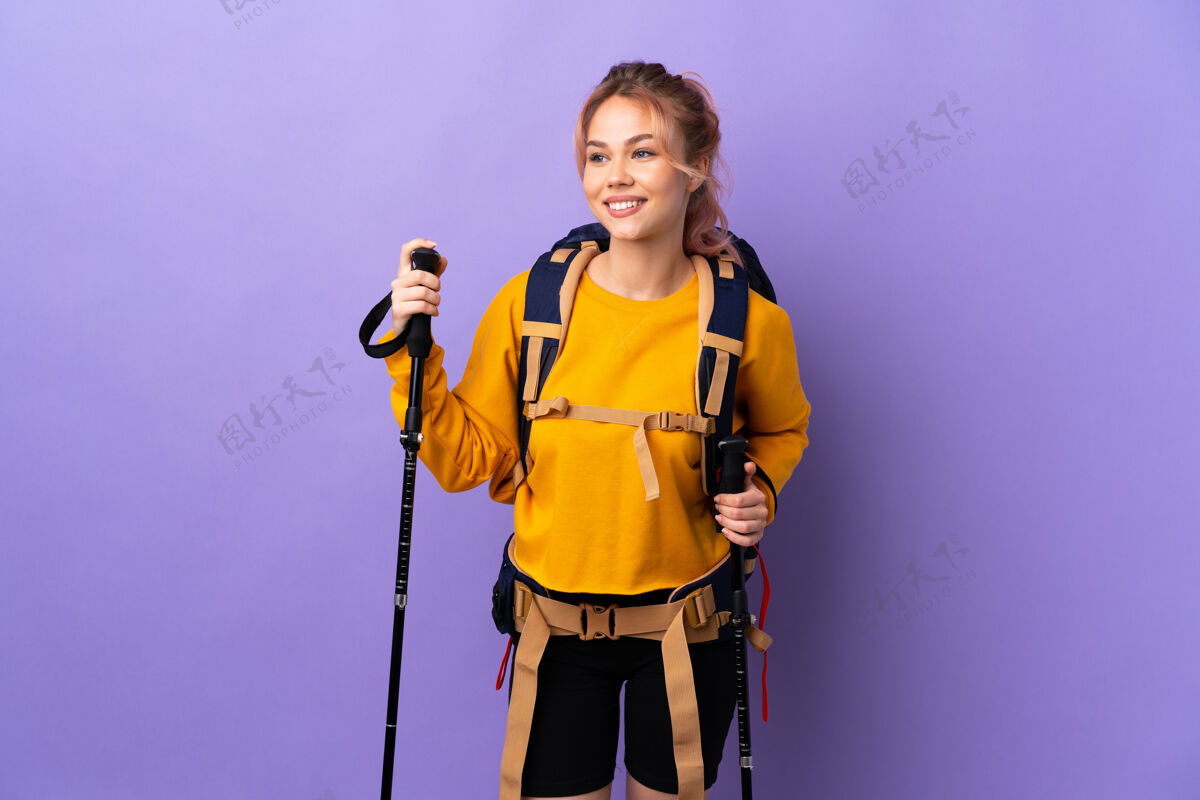 散步女孩背着背包和登山竿在孤立的紫色摆姿势 双臂放在臀部 面带微笑大笑自信脸