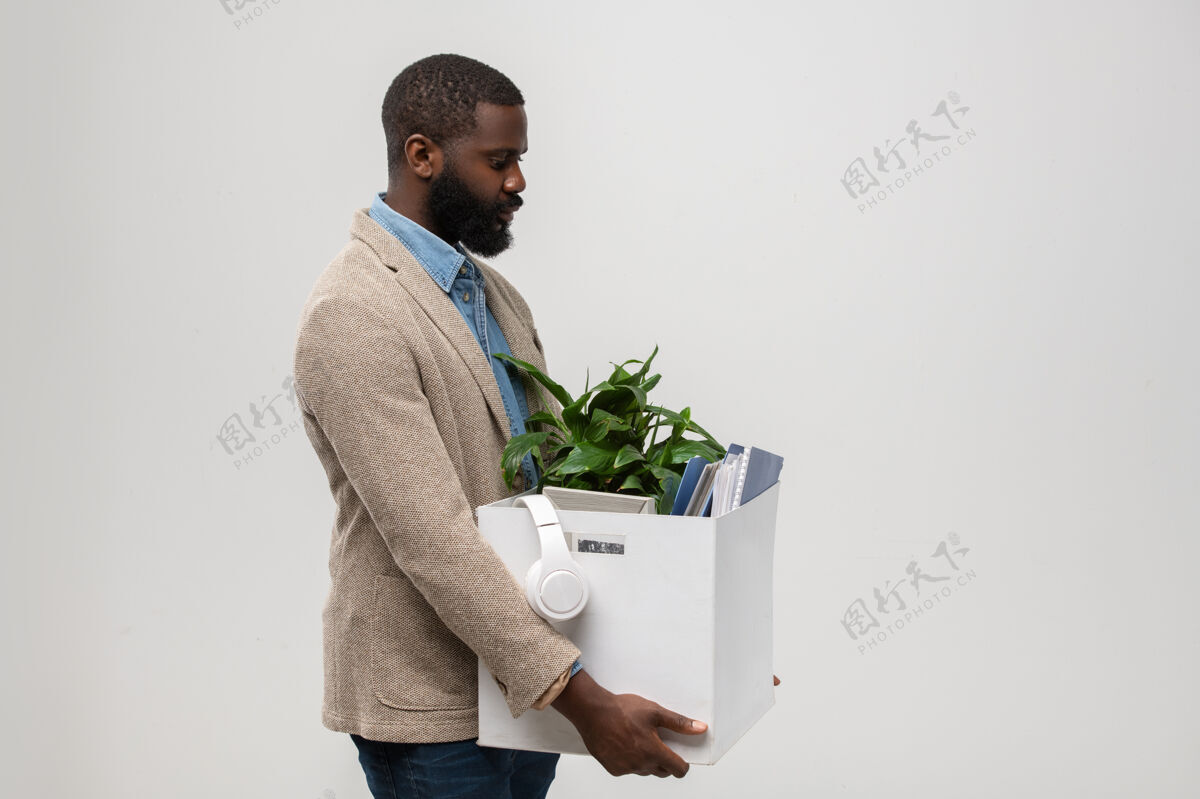 年轻人一位留着胡须的年轻美籍非洲裔办公室经理的侧视图 他穿着正装 看着带着耳机 绿色植物和其他用品的盒子室内专业职业成人