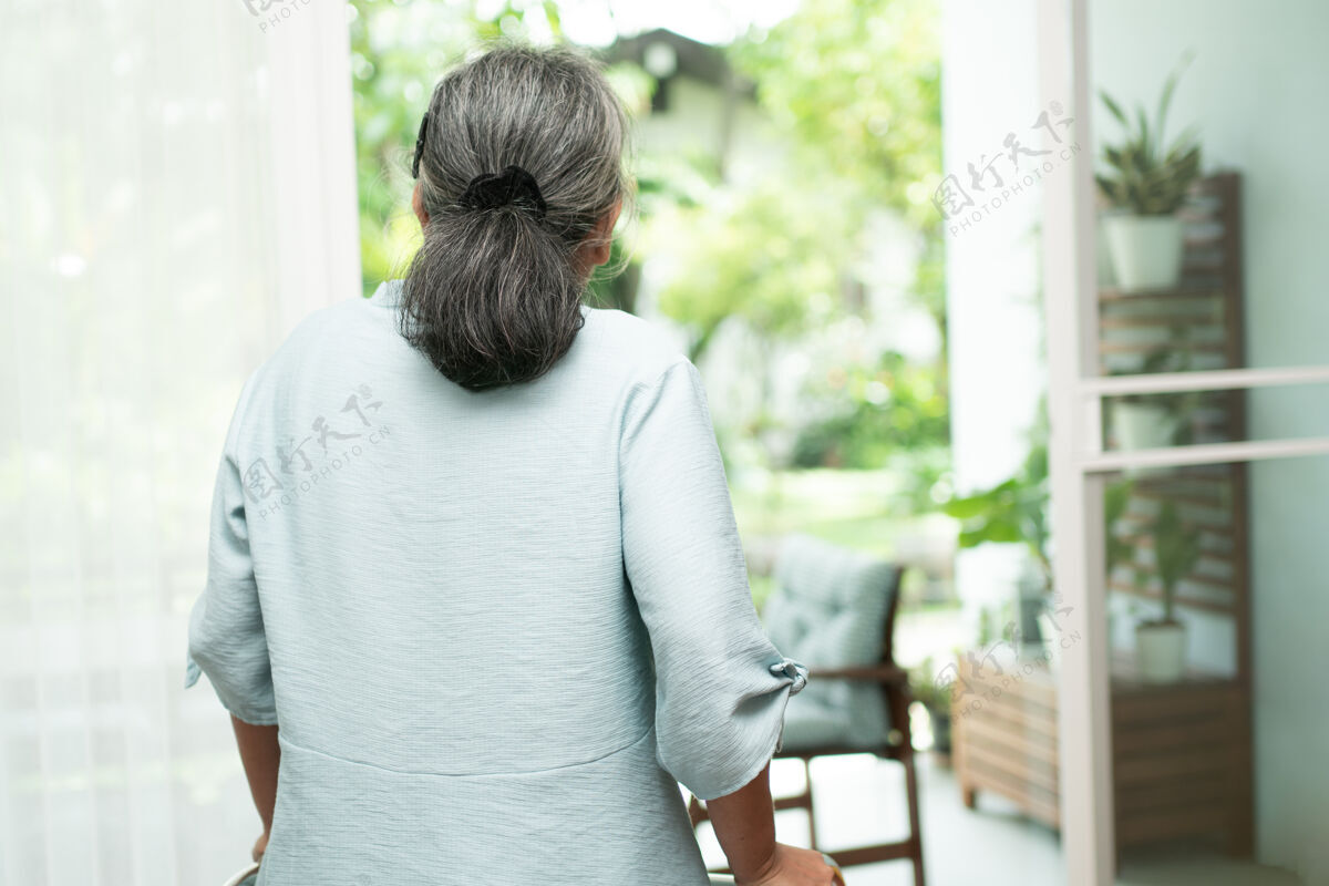 沉思一个悲伤的老妇人用助行器站在窗前 看着窗外 感到孤独人老人关心