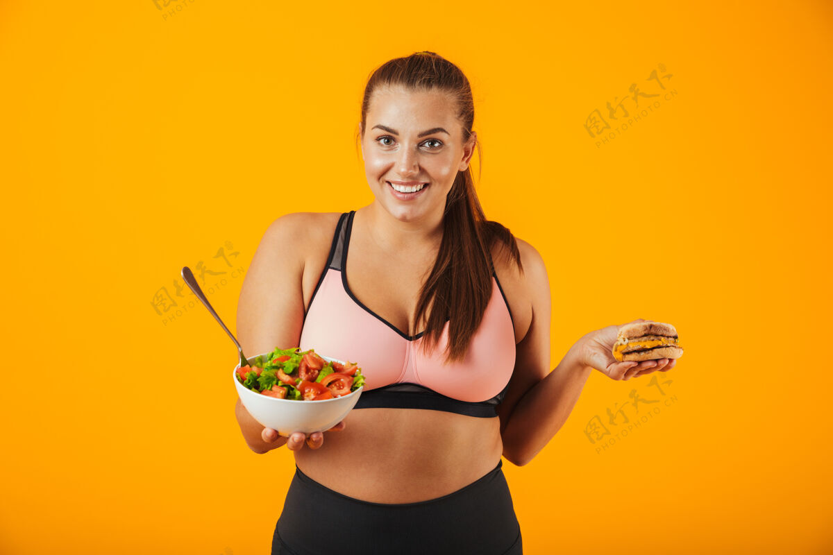 运动一个穿着运动服的快乐的超重健身妇女的肖像 孤立地站在黄色的墙上 手里拿着盛着沙拉和汉堡的碗蔬菜人体重