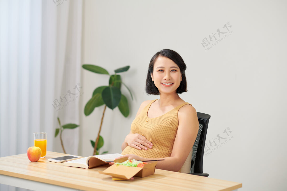 放松怀孕的年轻女子坐在客厅的桌子上使用数码平板电脑 吃早餐和工作房子年轻人女人