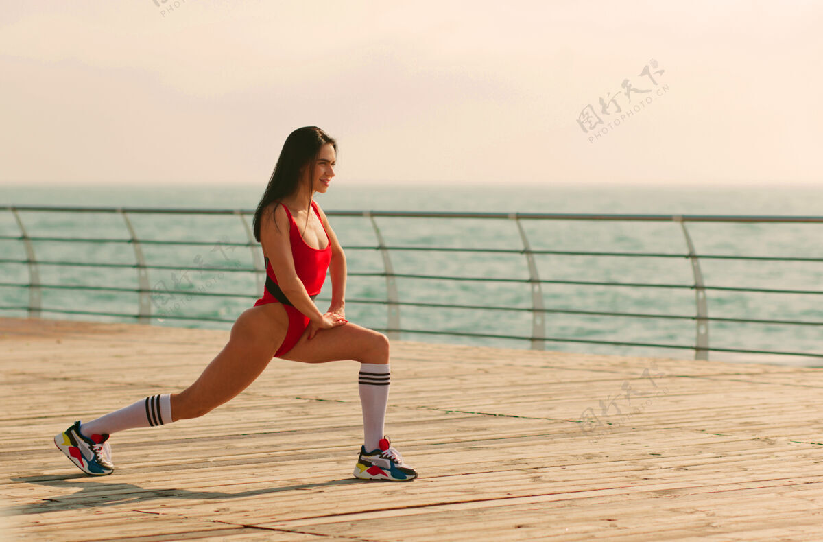 柔韧穿着红色运动紧身衣和运动鞋的年轻健美女士在沙滩上做有氧运动女人健身运动