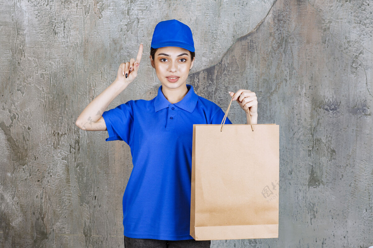 年轻穿着蓝色制服的女服务人员拿着一个纸板购物袋 想着有个主意外卖人体模型女售货员