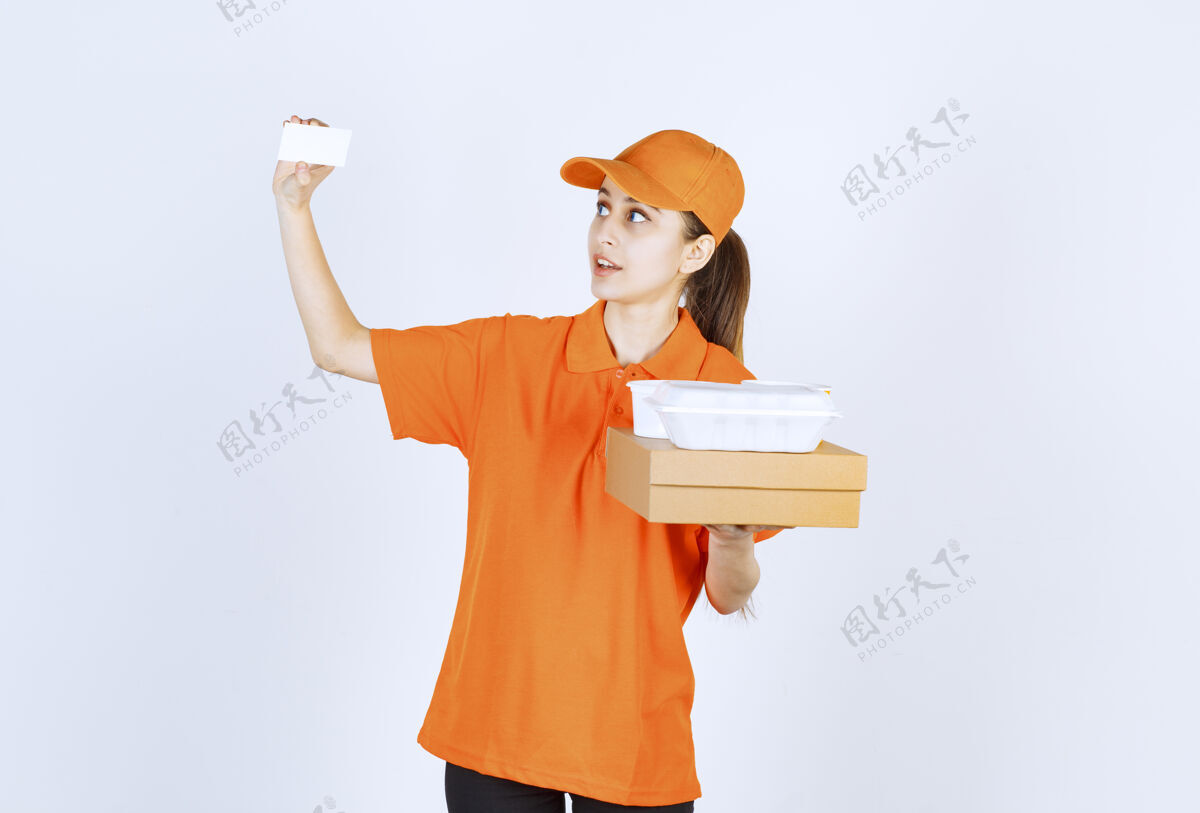 订单身着橙色制服的女快递员在出示名片时 手里拿着一个纸板箱和一个塑料外卖箱预订服装年轻人
