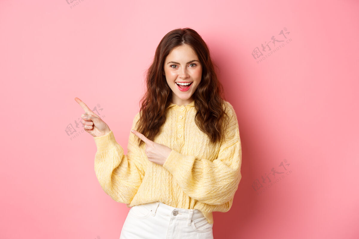 休闲自信快乐的女人 卷发 手指放在一边 在左边的版面上展示宣传片 兴奋地微笑着 站在粉色的墙上指向魅力女性