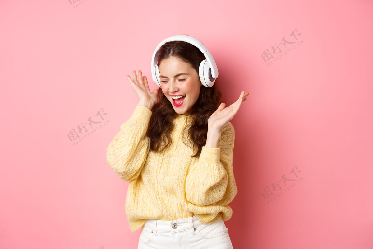 咧嘴笑快乐的女孩唱着最喜欢的歌 戴着无线耳机听音乐 微笑着跳舞 站在粉色的墙上微笑快乐欢呼