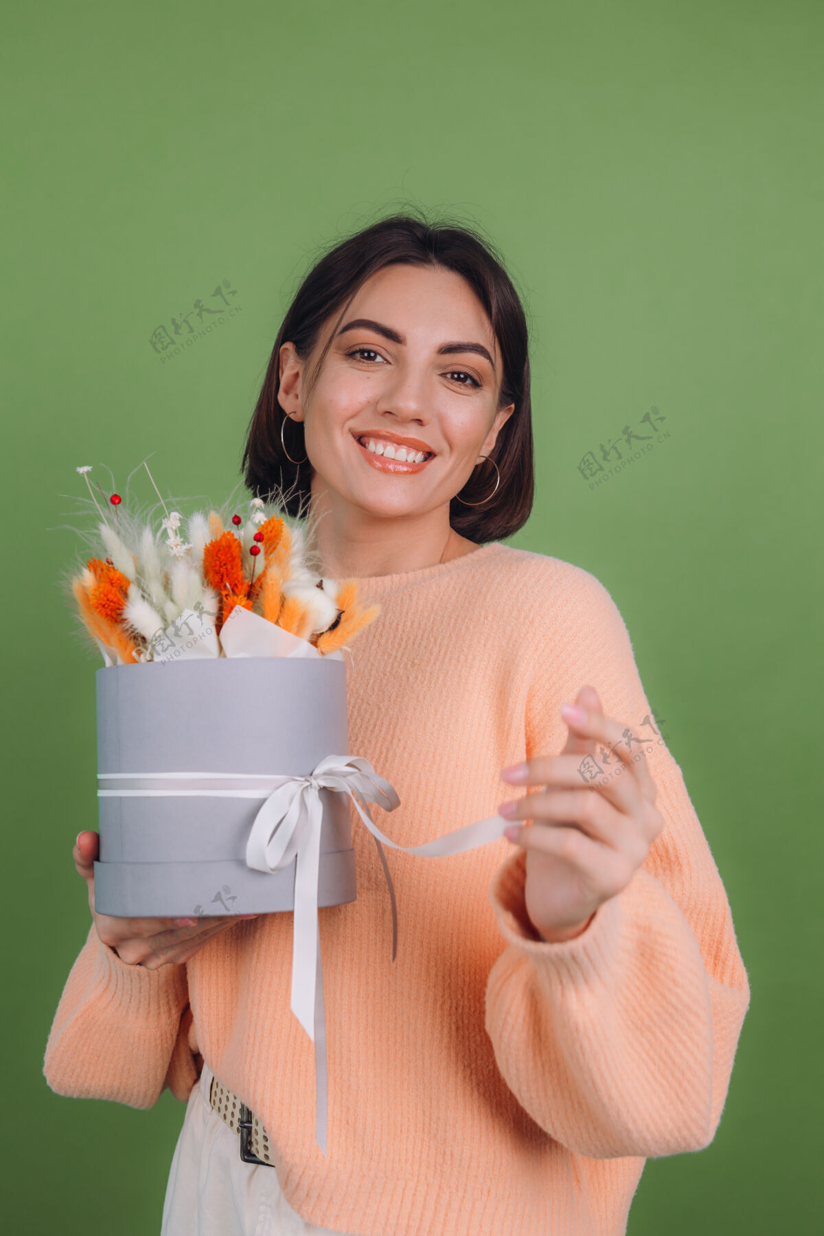 冷静年轻女子穿着休闲桃色毛衣隔离在绿橄榄墙上手持橙白色花盒组成的棉花 吉普赛拉小麦和拉古鲁斯作为礼物开心惊喜干花花束帽盒花