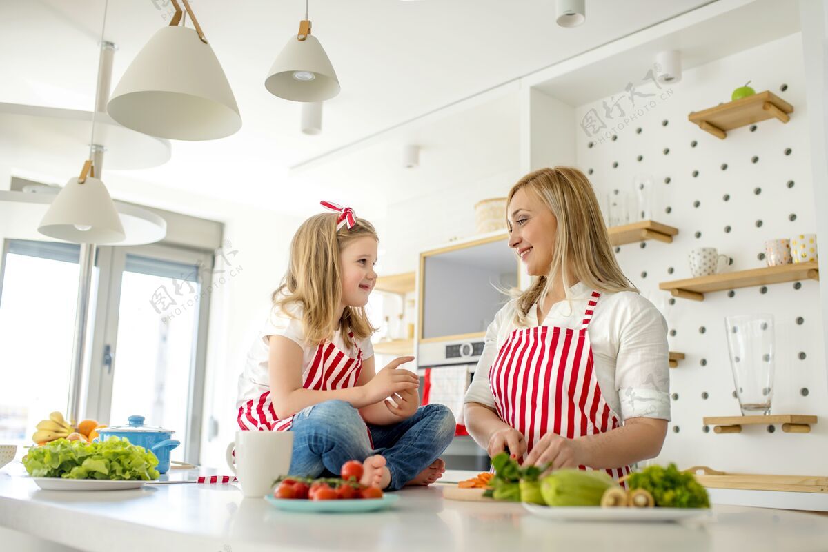 女性年轻的白人母亲在厨房里穿着相配的围裙在柜台上和女儿说话食物一起爱