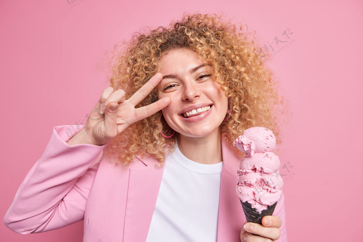 高兴积极漂亮的女人卷发吃巨大的冰淇淋在圆锥形华夫饼使和平姿态微笑广泛有乐趣在夏日穿着时髦的衣服隔离在粉红色的墙壁脸微笑女性