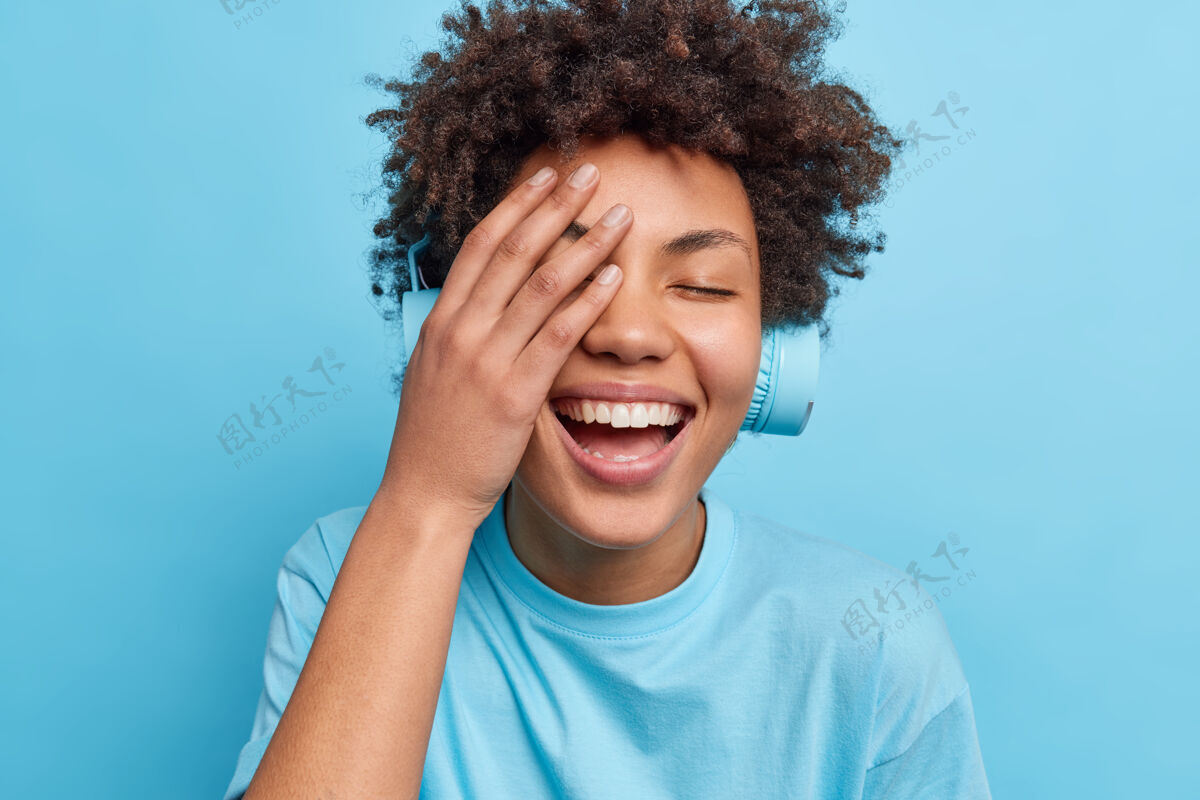 音频积极的咖喱头发的女人使脸掌心微笑愉快有无忧无虑的表情通过耳机听音频轨道穿着休闲t恤隔离在蓝色的墙壁上情感生活方式耳机单色黑发
