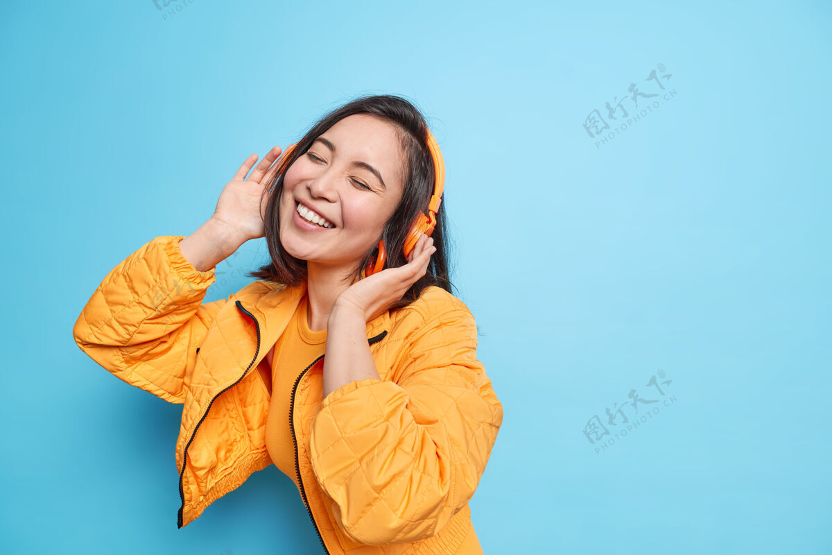 微笑年轻可爱的亚洲女人睁大眼睛 微笑 表情愉快 戴着耳机听音乐 穿着时髦的衣服 在蓝色的墙上欣赏最喜欢的歌曲可爱时尚人