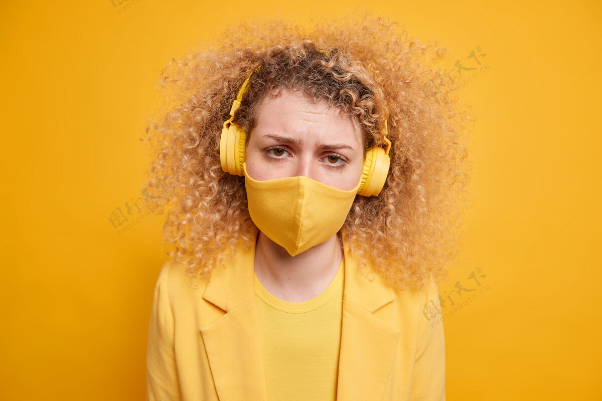 耳机失望不高兴的年轻女子戴着防护面具感到孤独独自呆在家里自我隔离通过无线立体声耳机听音乐对着黄色的墙壁摆姿势预防呆在家里旋律