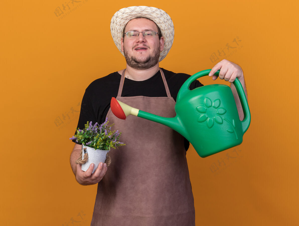 帽子自信的年轻男园丁戴着园艺帽和手套拿着浇水罐 花盆里的花被隔离在橙色的墙上年轻人花盆拿着