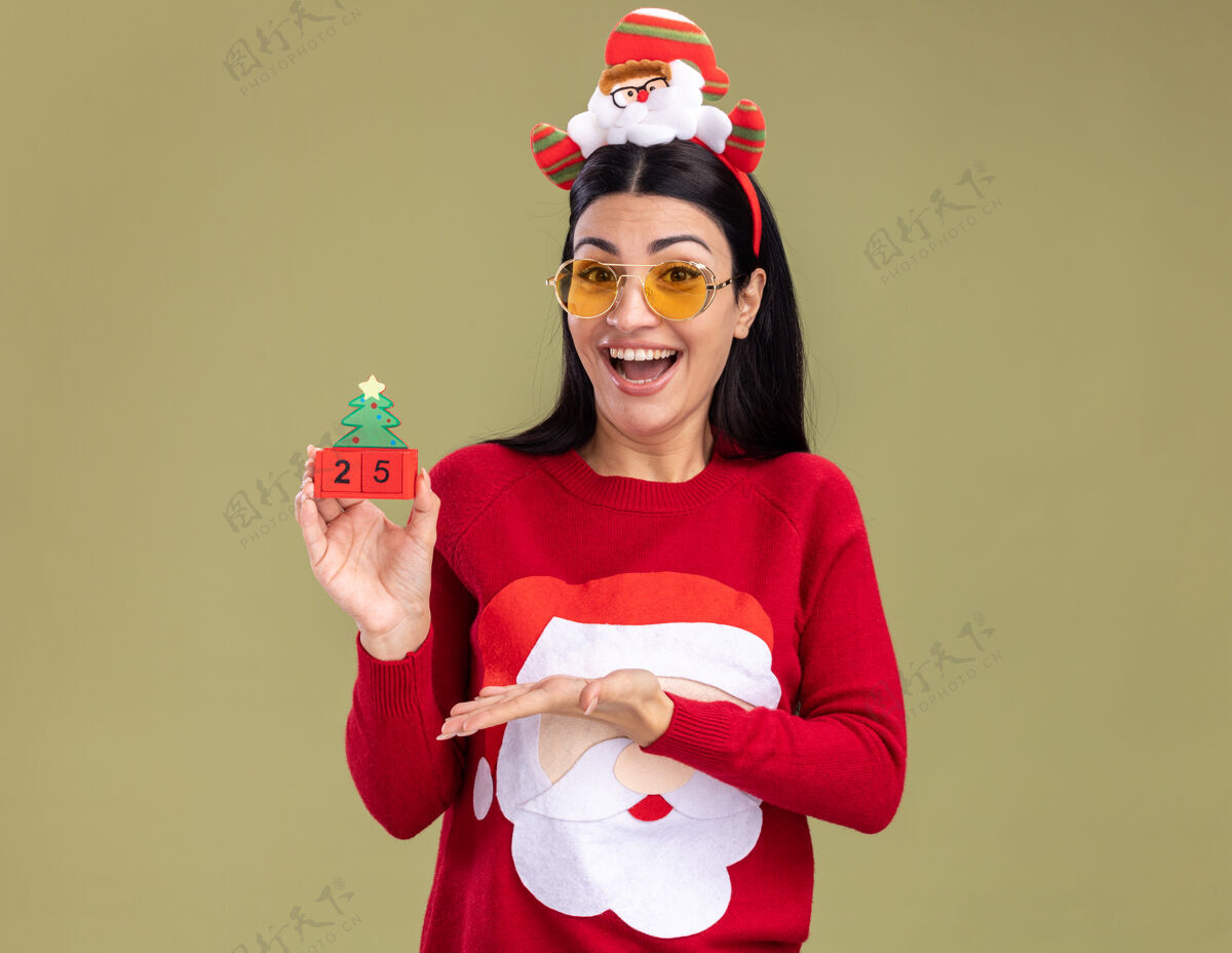 毛衣快乐的年轻白人女孩戴着圣诞老人的头带和带眼镜的毛衣拿着圣诞树玩具和日期指着它隔离在橄榄绿的墙上复制空间白种人女孩圣诞老人