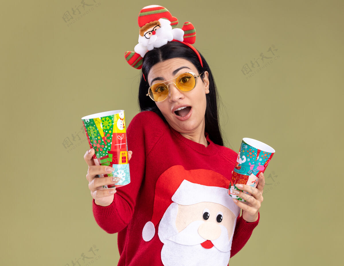 年轻令人印象深刻的年轻白人女孩戴着圣诞老人的头带和毛衣与眼镜举行塑料圣诞杯伸出一个橄榄绿的墙壁上孤立毛衣印象举行
