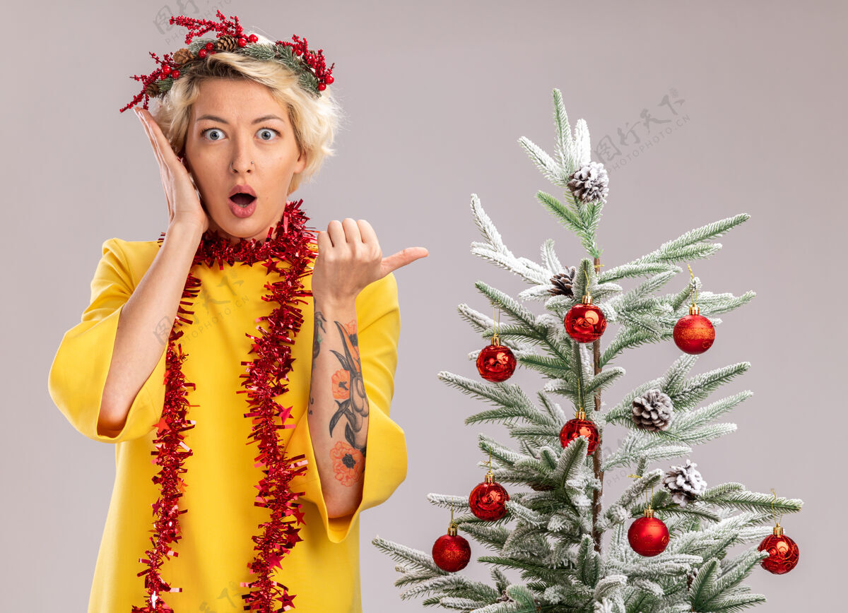 保持关心的金发女郎戴着圣诞花环 脖子上戴着金属丝花环 站在装饰好的圣诞树旁 看着手贴着脸 指着隔离在白色墙上的一面关心金属片壁板