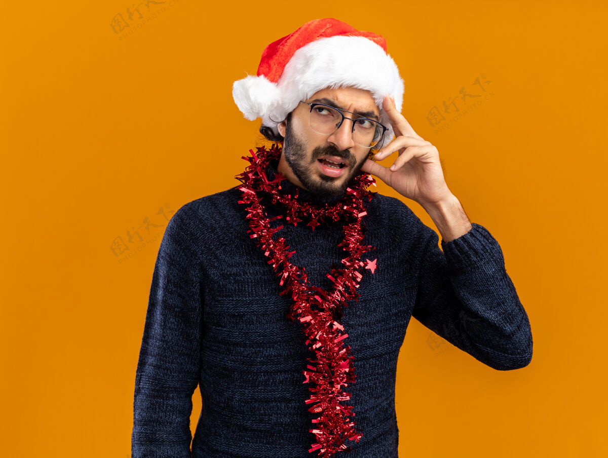 手一边想一边年轻帅哥戴着圣诞帽 脖子上戴着花环 手放在头上 隔离在橙色的墙上年轻脖子帅哥