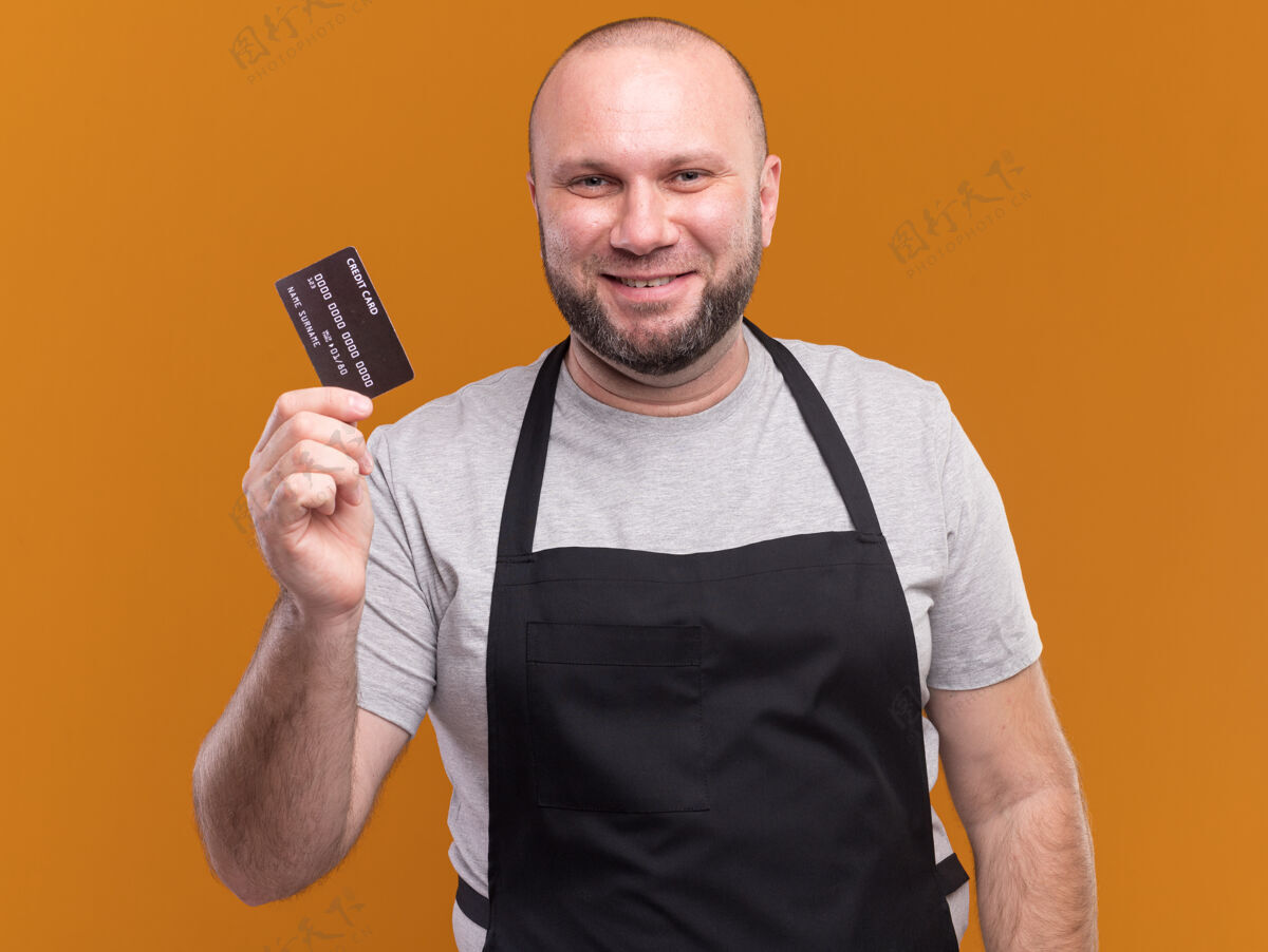 人高兴的中年男理发师穿着制服拿着信用卡隔离在橙色的墙上公民姿势持有