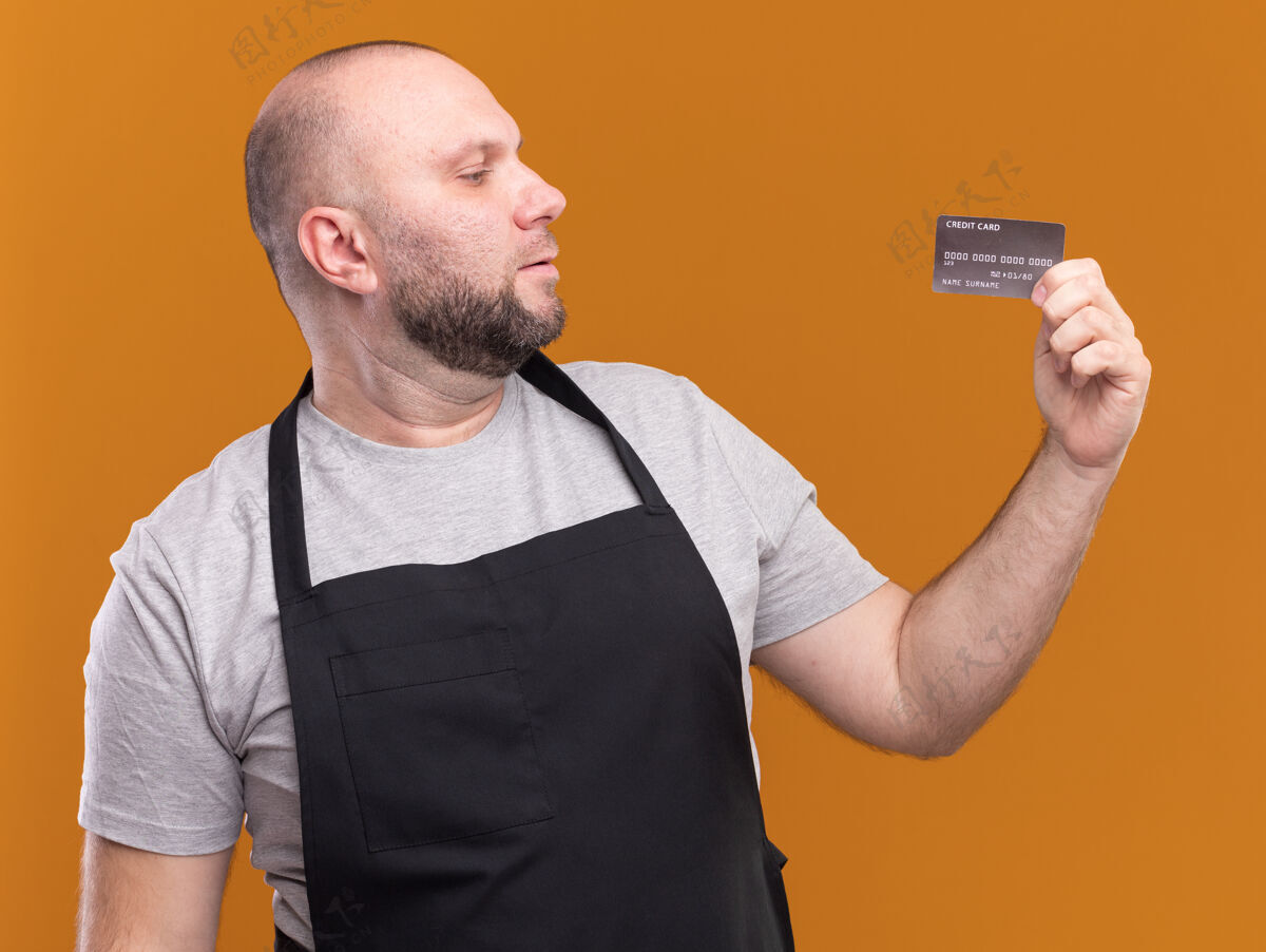 制服想着中年男理发师穿着制服拿着信用卡看着隔离在橙色墙上立场人人