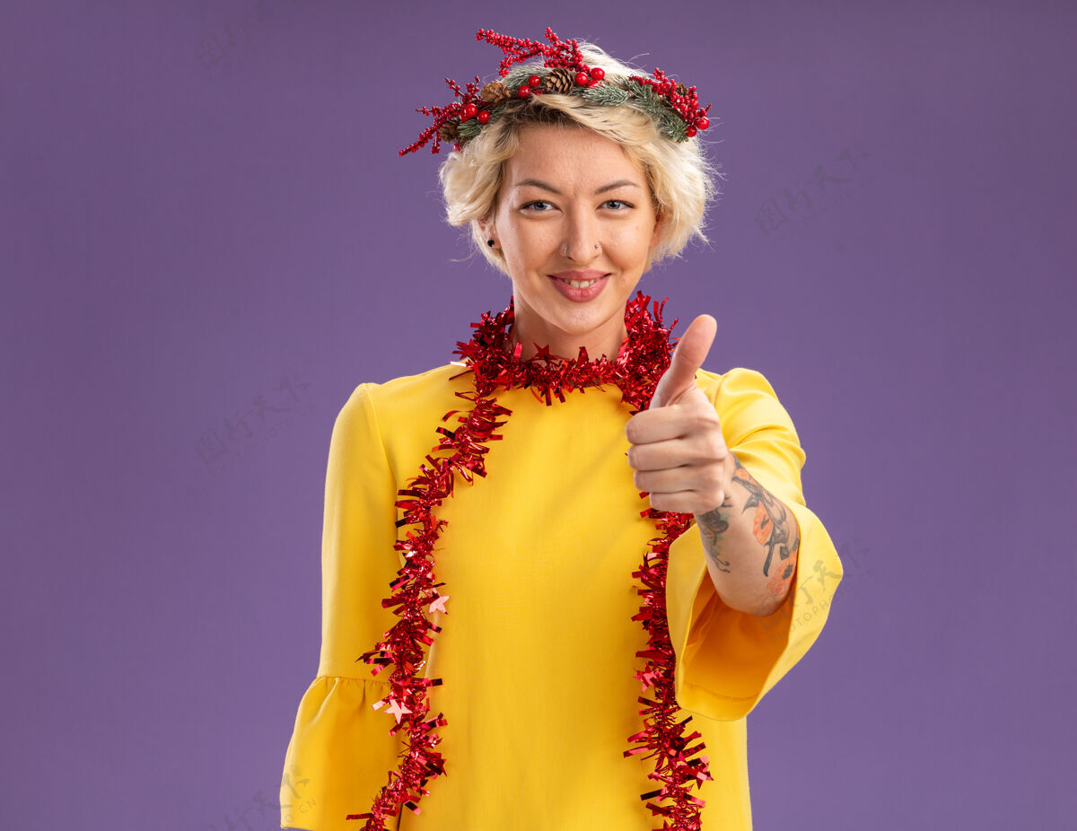 年轻年轻的金发女郎戴着圣诞花环 脖子上戴着金箔花环 看起来很高兴 在紫色的墙上孤立地竖起大拇指 留着复制空间头脖子花环