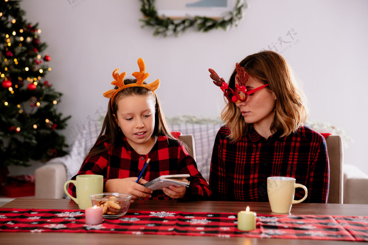桌子戴着驯鹿眼镜的妈妈高兴地看着拿着铅笔和笔记本的女儿坐在桌旁 在家里享受圣诞节的时光抱着女儿坐着