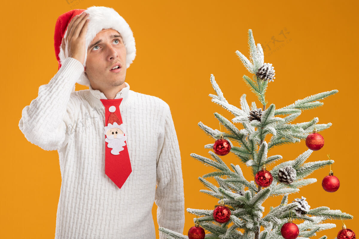 圣诞老人遗憾的是 年轻帅哥戴着圣诞帽 打着圣诞老人的领带 站在装饰好的圣诞树旁 手放在头上 孤零零地看着橙色的墙上后悔圣诞老人穿