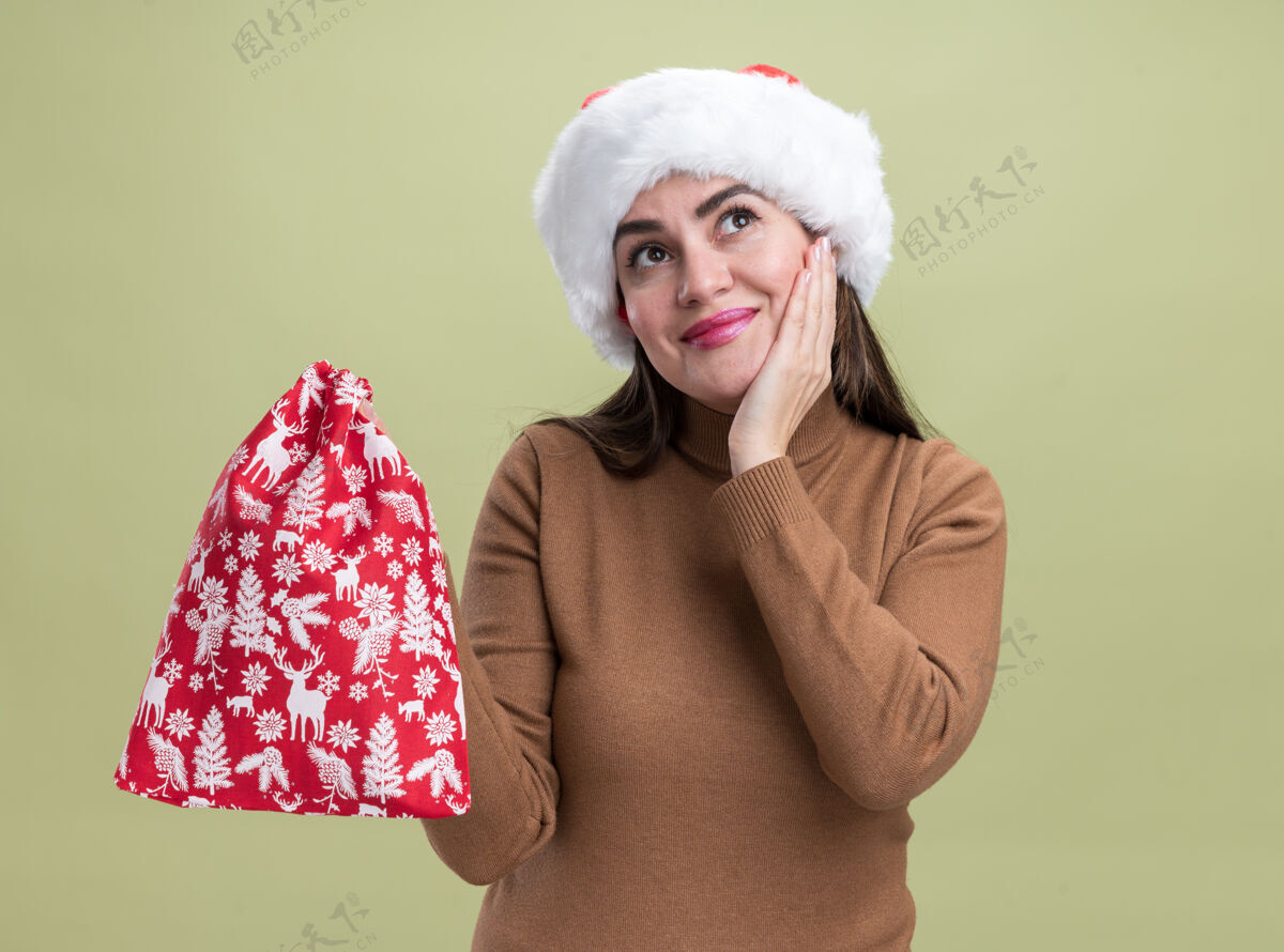 请高兴的年轻漂亮女孩戴着圣诞帽拿着圣诞包把手放在脸颊上隔离在橄榄绿的墙上包脸颊帽子