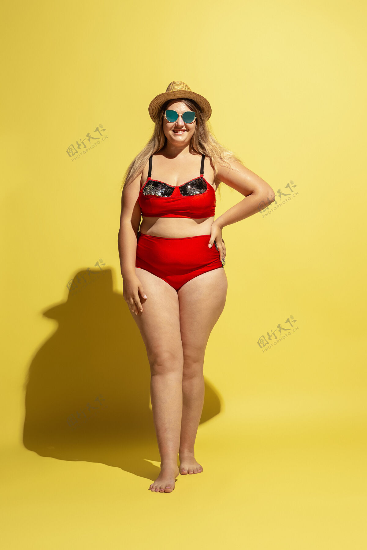 假日年轻的白人大码女模特在黄墙上准备度假穿着红色泳衣 戴着帽子和太阳镜的女人假期超重举行
