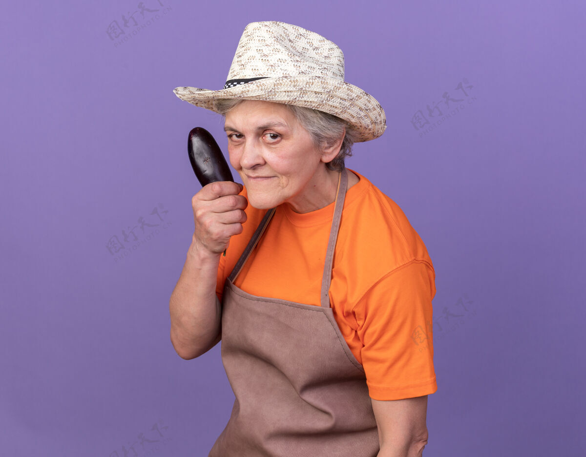 紫色自信的上了年纪的女园丁戴着园艺帽拿着茄子穿女花园