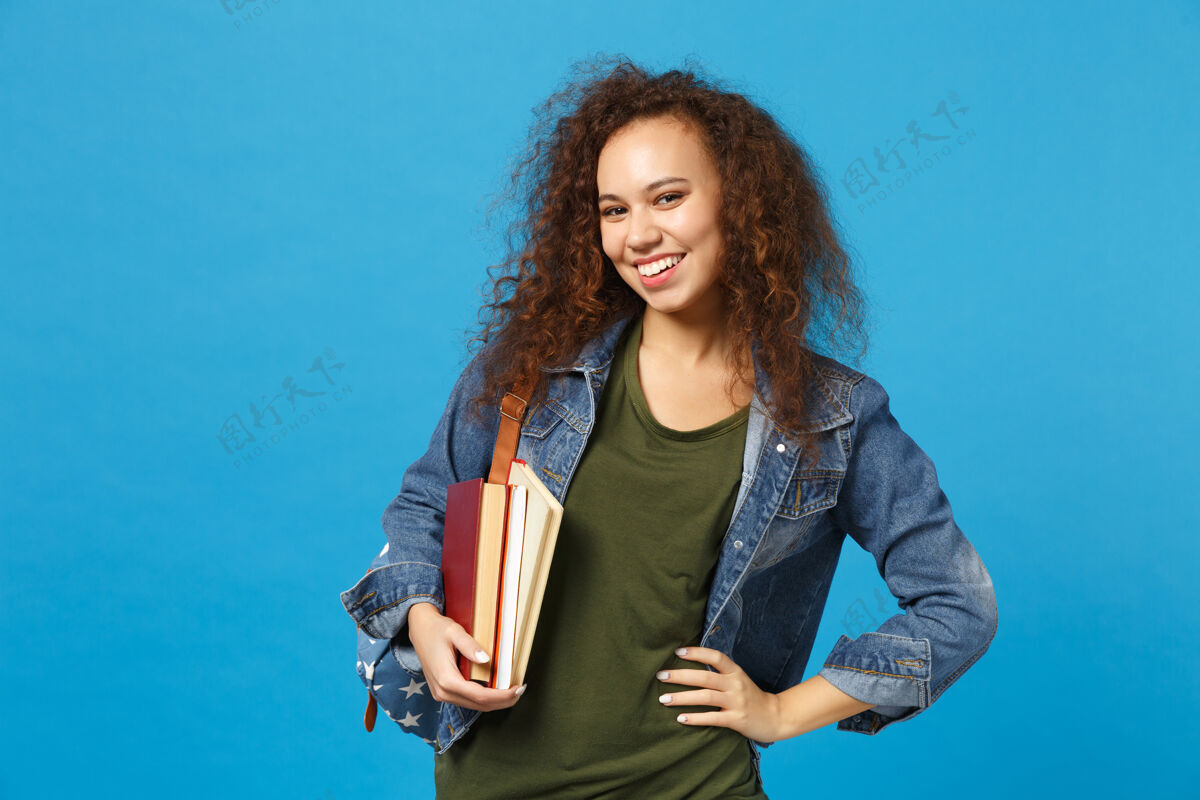 背包穿着牛仔服 背着书包的年轻女学生把书孤立地放在蓝色的墙上学习牛仔裤站