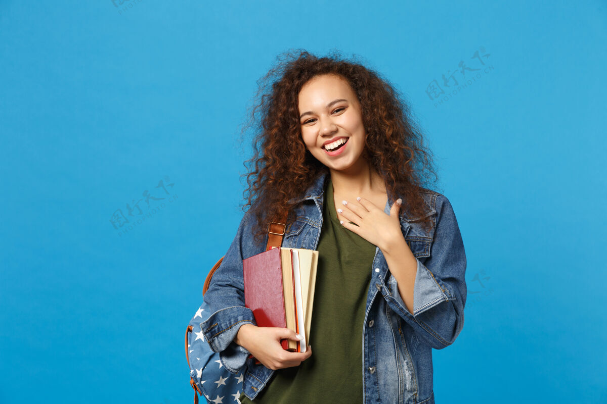学生穿着牛仔服 背着书包的年轻女学生把书孤立地放在蓝色的墙上青年采取聪明