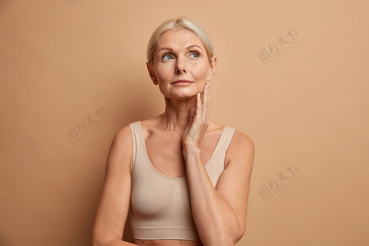 老年人女人在涂上浓缩的抗衰老面霜后触摸皮肤 表情若有所思 穿着棕色的不规则剪裁上衣面部柔嫩皱纹